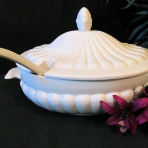 May include: White ceramic serving bowl with a lid and a matching ladle. The bowl has a fluted design and the lid has a knob on top. The bowl is sitting on a black surface.