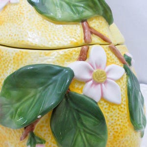 May include: A ceramic jar shaped like a yellow lemon with a textured rind. The jar features three-dimensional green leaves, brown stems, and white blossoms with yellow centers attached to the exterior surface. The lid is topped with a stem.