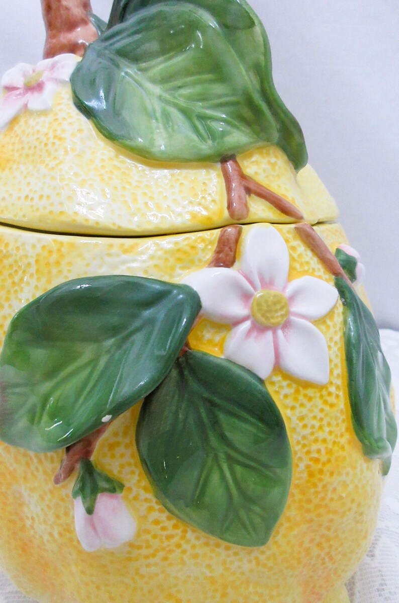 May include: A ceramic jar shaped like a yellow lemon with a textured rind. The jar features three-dimensional green leaves, brown stems, and white blossoms with yellow centers attached to the exterior surface. The lid is topped with a stem.