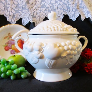以下が含まれることがあります： 白いセラミック製のスープ皿で、蓋付き、エンボス加工されたフルーツのデザインが施されています。スープ皿には2つのハンドルがあり、台座の上に置かれています。フルーツの装飾皿と青リンゴが近くにあります。