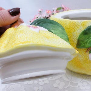 May include: A ceramic canister with a lid, featuring a textured yellow lemon-shaped exterior. The design includes three-dimensional green leaves and small white and pink blossoms attached to the surface of both the lid and the main container.