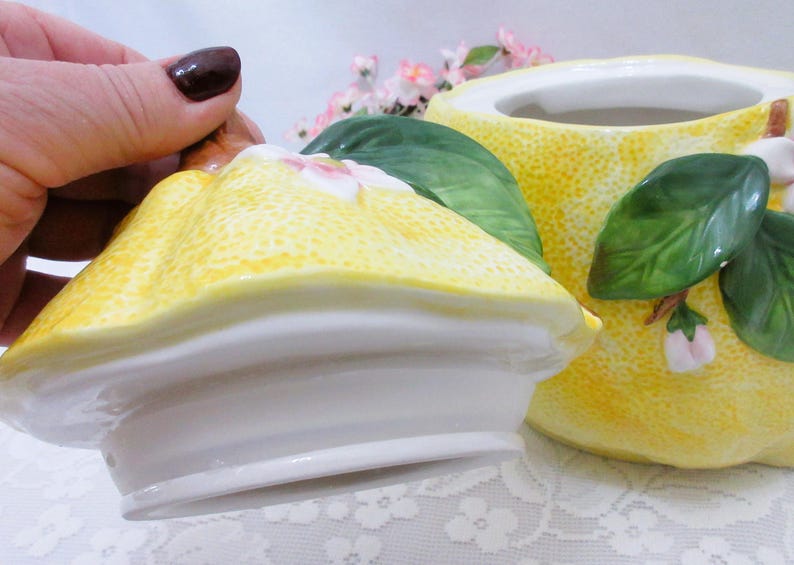 May include: A ceramic canister with a lid, featuring a textured yellow lemon-shaped exterior. The design includes three-dimensional green leaves and small white and pink blossoms attached to the surface of both the lid and the main container.