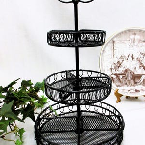 May include: A black, three-tiered serving stand with a decorative floral top. The stand features three circular wire mesh trays, perfect for displaying desserts or small items. The stand is made of metal and has a classic design.