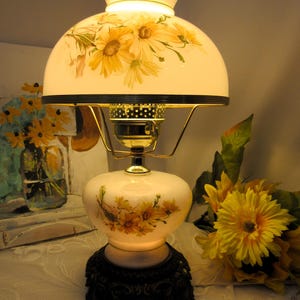 May include: A table lamp with a milk glass shade and base, both decorated with yellow daisy floral patterns. The lamp features a brass-toned metal frame, a scalloped top edge on the shade, and a dark, ornate, multi-footed base stand.