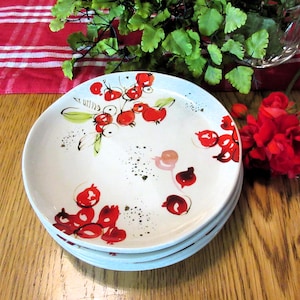 Könnte beinhalten: Ein Stapel weißer Keramikteller mit einem handgemalten Design aus roten Beeren und grünen Blättern. Die Teller haben einen leicht gewellten Rand und sind auf einer Holzoberfläche angeordnet. Das Design umfasst schwarze Sprenkel und einige rosa Akzente.