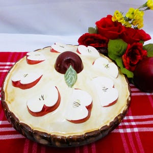 May include: A ceramic apple pie dish with a brown crust and a cream-colored filling, decorated with apple slices. A whole apple and red roses with yellow accents sit nearby on a red and white checkered tablecloth.