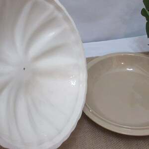 以下が含まれることがあります： オフホワイトのセラミック製サービングディッシュ。蓋には渦巻き模様が施されています。皿はベージュ色で丸く、縁が少し盛り上がっています。蓋は開いています。