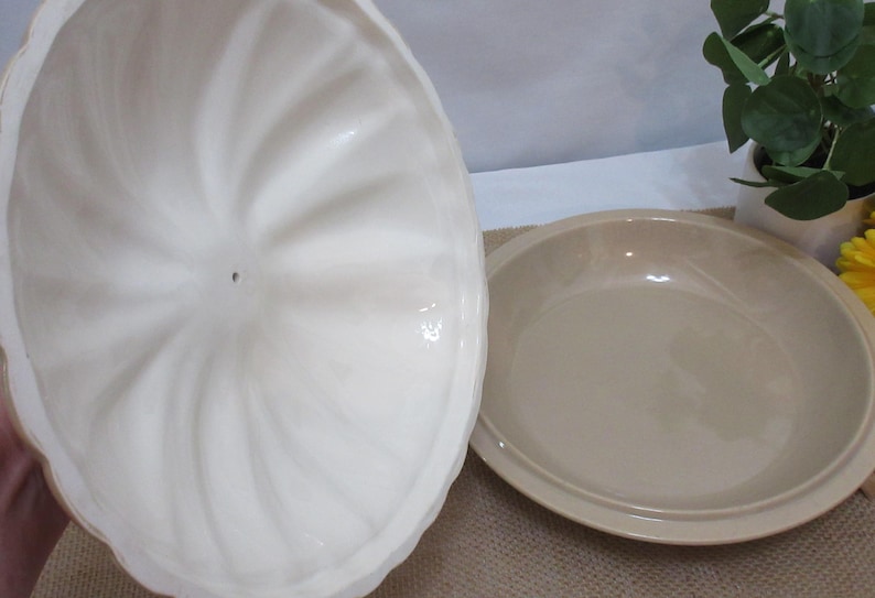 以下が含まれることがあります： オフホワイトのセラミック製サービングディッシュ。蓋には渦巻き模様が施されています。皿はベージュ色で丸く、縁が少し盛り上がっています。蓋は開いています。