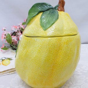 May include: A ceramic jar shaped like a large yellow lemon with a textured rind. The lid features a brown stem and two green leaves attached to the top. The surface of the jar has a bumpy, realistic lemon peel texture and a glossy finish.