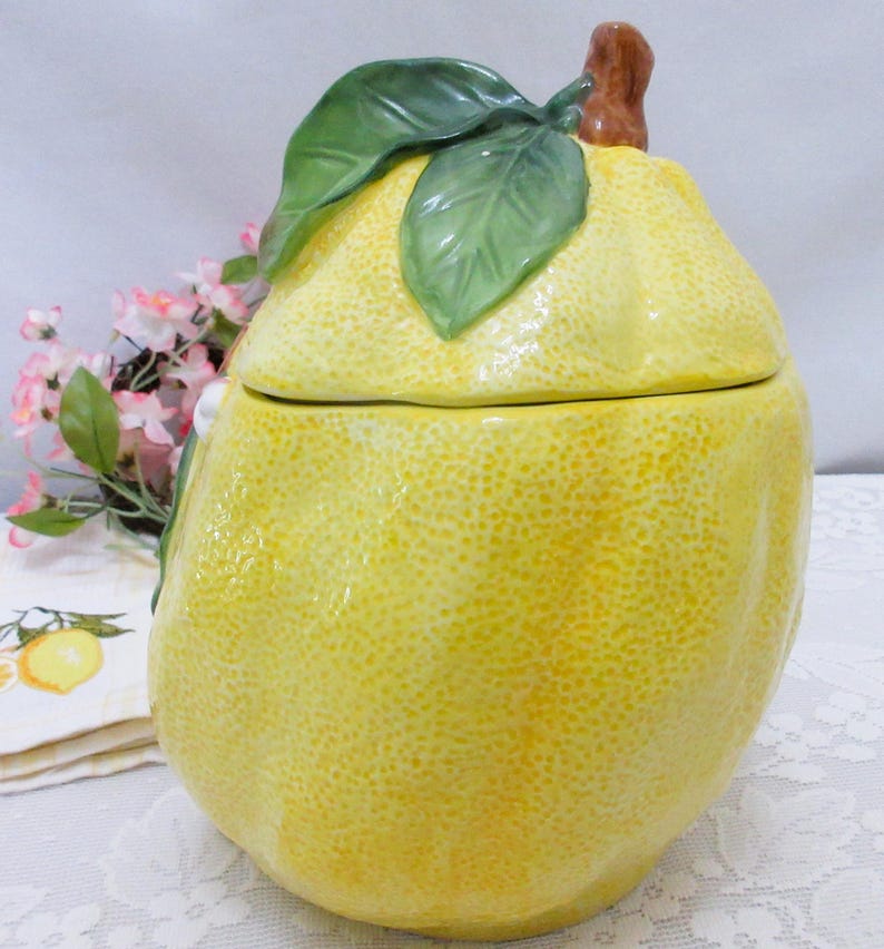 May include: A ceramic jar shaped like a large yellow lemon with a textured rind. The lid features a brown stem and two green leaves attached to the top. The surface of the jar has a bumpy, realistic lemon peel texture and a glossy finish.