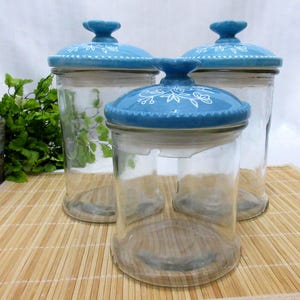 May include: Three clear glass jars with blue lids. The lids have a floral design in white. The jars are on a bamboo mat with greenery in the background. The jars are empty.