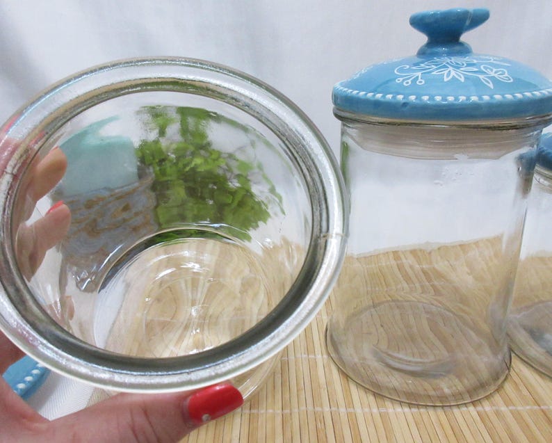 May include: Two clear glass jars with blue and white lids. The lids have a floral design. The jars are empty.
