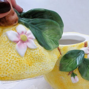 May include: A ceramic sugar bowl shaped like a lemon with a textured yellow surface, featuring sculpted green leaves and white flowers with pink centers. The lid is removable and includes a brown stem handle, while the base is a matching lemon.
