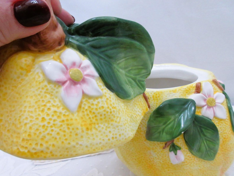 May include: A ceramic sugar bowl shaped like a lemon with a textured yellow surface, featuring sculpted green leaves and white flowers with pink centers. The lid is removable and includes a brown stem handle, while the base is a matching lemon.