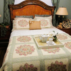 May include: A bedroom scene featuring a wooden headboard, floral patterned bedding, and a white tray with a teapot and cup. The quilt has large floral blocks in pink and cream, with a green border. Pillows and curtains complete the cozy, inviting aesthetic.