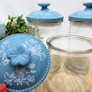 May include: Three glass jars with blue and white ceramic lids. The lids feature a floral design. The jars are empty.