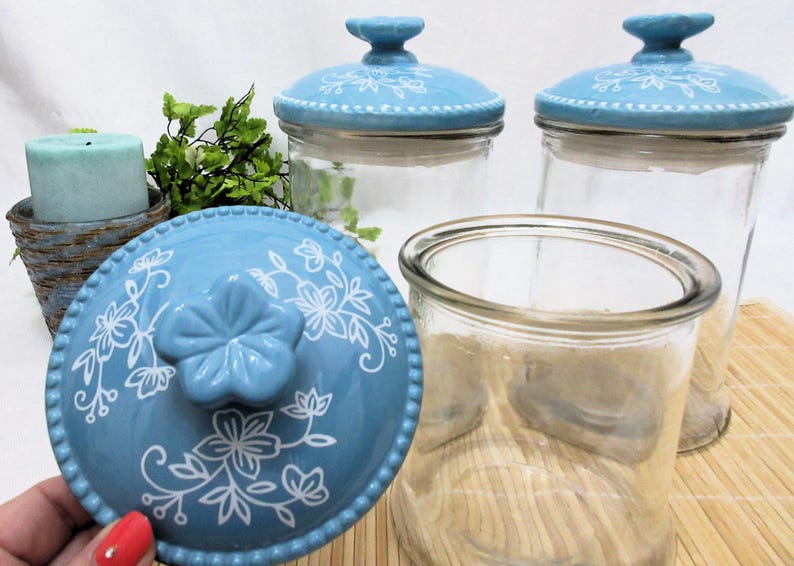 May include: Three glass jars with blue and white ceramic lids. The lids feature a floral design. The jars are empty.