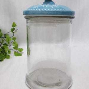 May include: A clear glass jar with a blue ceramic lid. The lid has a white floral design and a small butterfly knob.
