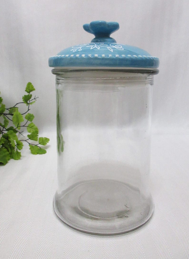 May include: A clear glass jar with a blue ceramic lid. The lid has a white floral design and a small butterfly knob.
