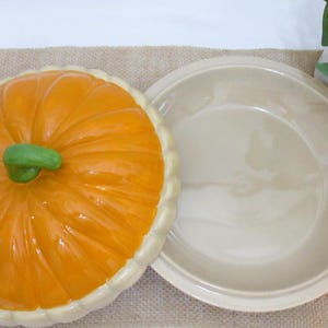 以下が含まれることがあります： 緑色の茎とクリーム色の縁が付いた、オレンジ色のセラミックカボチャ型パイ皿。蓋はパイ皿のベースの隣にあります。パイ皿はジュートの表面にあります。秋をテーマにした装飾品。
