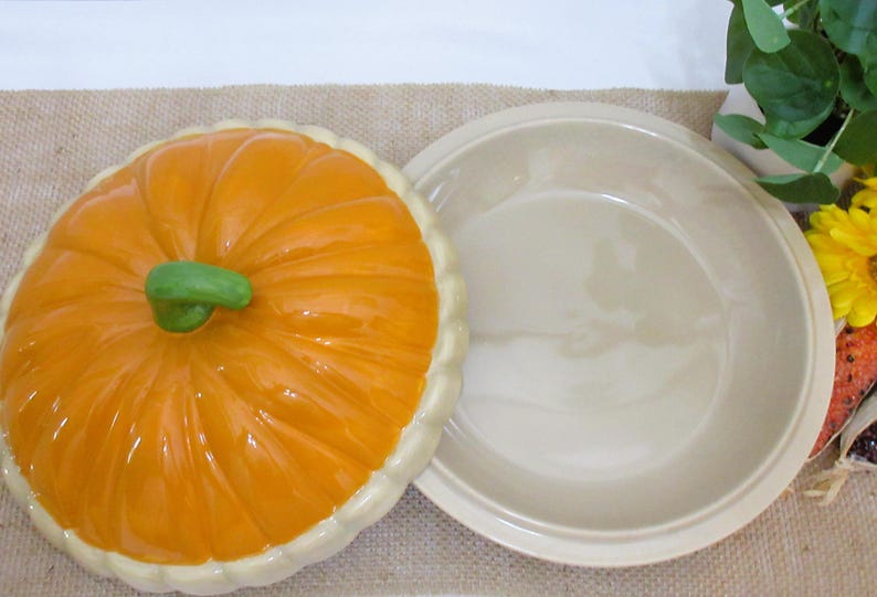 以下が含まれることがあります： 緑色の茎とクリーム色の縁が付いた、オレンジ色のセラミックカボチャ型パイ皿。蓋はパイ皿のベースの隣にあります。パイ皿はジュートの表面にあります。秋をテーマにした装飾品。