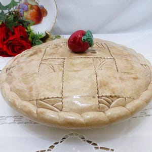 Pode incluir: Um prato de cerâmica redondo para tortas com tampa, apresentando uma textura de crosta entrelaçada em tom bege e acabamento brilhante. A tampa é rematada por um pequeno puxador tridimensional em forma de maçã vermelha com uma folha verde.