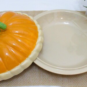 以下が含まれることがあります： 蓋付きのセラミック製カボチャ型サービングディッシュ。蓋はオレンジ色で、緑色の茎とクリーム色の縁が付いています。お皿はベージュ色で丸いです。秋の季節に食べ物を提供するのに適しています。