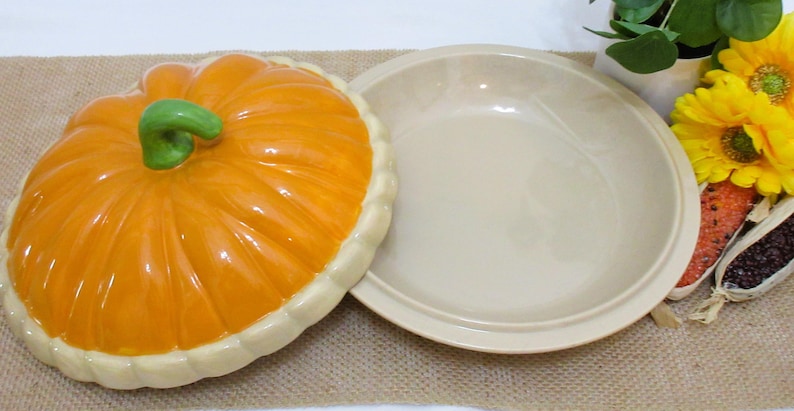 以下が含まれることがあります： 蓋付きのセラミック製カボチャ型サービングディッシュ。蓋はオレンジ色で、緑色の茎とクリーム色の縁が付いています。お皿はベージュ色で丸いです。秋の季節に食べ物を提供するのに適しています。