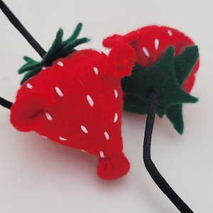 May include: Two felt strawberry hair clips with black elastic bands. The strawberries are red with white seeds and green leaves.