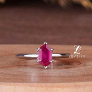 May include: A silver ring featuring a vibrant, hexagonal ruby gemstone. The ruby is a deep pink color and is the focal point of the ring. The ring is set on a wooden surface, with a blurred background.
