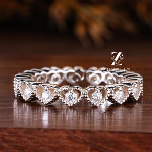 Op de afbeelding: Een zilveren eternity ring met hartvormige edelstenen. De ring ligt op een donker houten oppervlak, met een vage achtergrond van bloemen en bladeren. Het ontwerp van de ring toont een doorlopend patroon van hartvormige stenen.