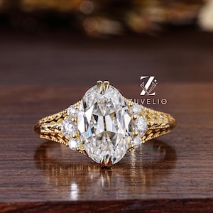 May include: A gold-colored ring with a large, oval-shaped, clear gemstone as the centerpiece. The ring has smaller, round gemstones on either side of the main stone. The band features intricate filigree detailing. The ring is displayed on a dark wooden surface.