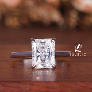 May include: A radiant-cut diamond engagement ring with a silver band. The large, rectangular diamond is the focal point, set on a simple, elegant band. The ring is displayed on a dark wooden surface, with the brand name "ZUVLIO" visible.
