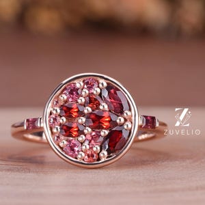 May include: A rose gold ring featuring a circular design with a cluster of red and pink gemstones. The ring has a delicate band and is set on a wooden surface. The ring is a piece of jewelry.