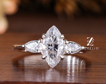 May include: A marquise-cut diamond engagement ring with two pear-shaped side stones. The ring is set in a silver band and sits on a wooden surface. The ring is a classic design, perfect for a proposal.