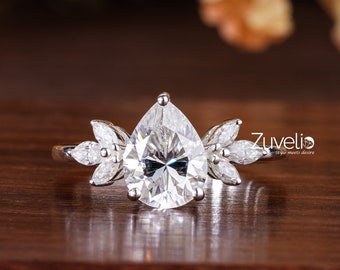 May include: A pear-shaped diamond engagement ring with a silver band and smaller diamonds on the sides. The ring is set on a wooden surface.