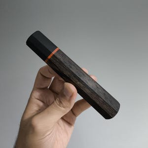 May include: A dark wooden pen holder with a black cap and an orange accent band. The pen holder is held at an angle against a grey background. The wood grain is visible on the pen holder.