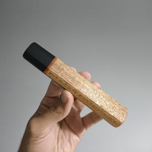 May include: A wooden knife handle with a black end and a thin orange accent. The handle is held in a hand and has a geometric design. The wood grain is visible.