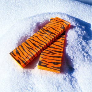 Op de afbeelding: Twee rechthoekige oranje en zwart gestreepte objecten liggen op witte sneeuw. De objecten hebben een heldere oranje basiskleur met zwarte strepen, die het patroon van een tijger nabootsen. Het zijn waarschijnlijk decoratieve of functionele items.