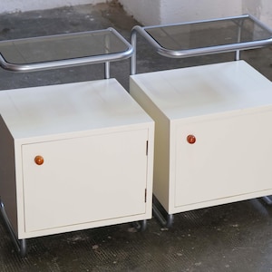 May include: A pair of off-white bedside cabinets featuring a single door with a small orange knob, supported by a tubular silver metal frame that extends upward to form a shelf with a dark tinted glass top surface for each unit.