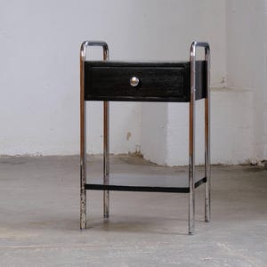 Könnte beinhalten: Ein schwarzer Nachttisch mit einer Schublade und einem silbernen Knopf. Der Nachttisch hat ein verchromtes Gestell und ein unteres Regal. Das Design ist eine Mischung aus modernem und Vintage-Stil. Der Tisch ist etwa 61 cm hoch.