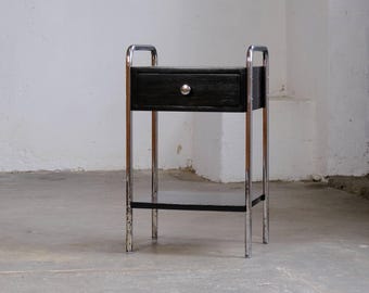 Könnte beinhalten: Ein schwarzer Nachttisch mit einer Schublade und einem silbernen Knopf. Der Nachttisch hat ein verchromtes Gestell und ein unteres Regal. Das Design ist eine Mischung aus modernem und Vintage-Stil. Der Tisch ist etwa 61 cm hoch.