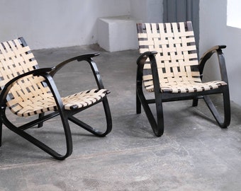 May include: Two black wooden chairs with woven beige and black seats. The chairs have a curved back and arms and are set on a concrete floor.