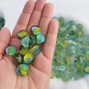 May include: A hand holding a pile of green, blue and yellow marbles. The marbles are in a variety of patterns. There are more marbles in the background.
