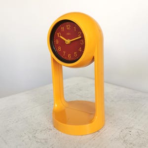 May include: A retro yellow alarm clock with a red clock face. The clock has a rounded top and a rectangular base with a curved handle. The clock face displays the numbers 1 to 12 in a white font.