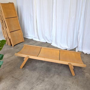 Op de afbeelding: Een houten opvouwbare strandstoel met een lichtbruine canvas zitting. De stoel staat in een ontspannen positie, met de poten uitgeschoven. Een andere stoel is opgevouwen en leunt tegen een muur. Witte gordijnen en een plant in pot op de achtergrond.