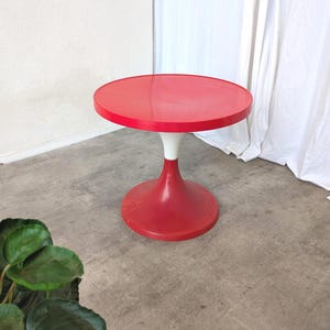 Puede incluir: Una mesa auxiliar roja y blanca con forma de tulipán. La mesa tiene una tapa redonda y una base acampanada. La mesa está hecha de un material brillante. La mesa tiene aproximadamente 51 cm de diámetro y 51 cm de altura.