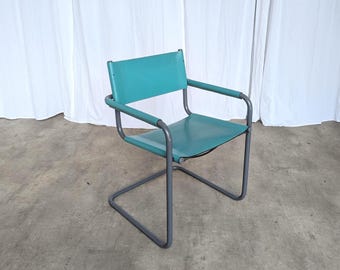 May include: A cantilever chair featuring a dark gray tubular metal frame and a teal seat and backrest. The chair has two armrests covered in matching teal material, and the seat and back are constructed from a smooth, solid teal panel.