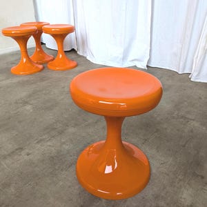 May include: Four orange, mid-century modern style stools. The stools have a round seat, a flared base, and a central stem. The stools are made of a glossy, solid orange material. The stools are on a concrete floor.