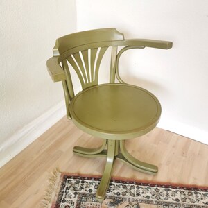 May include: An olive green vintage swivel chair with a curved back and armrest. The chair has a round seat and a five-point base. The chair is made of wood and has a classic design. The chair is placed on a light wood floor.